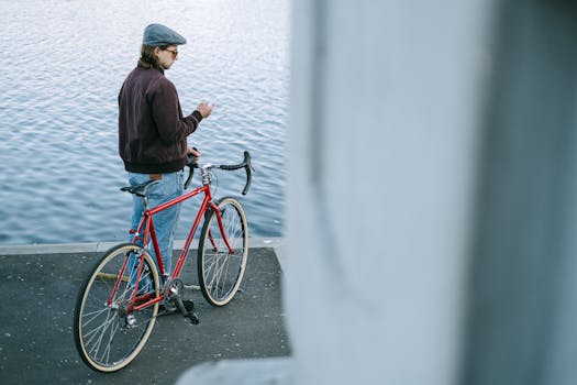 A man standing by a lake, looking at his smartphone while holding a red bicycle.