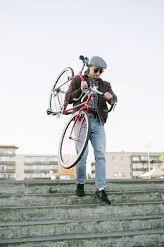 A stylish man in casual attire carrying a bicycle down outdoor concrete steps.
