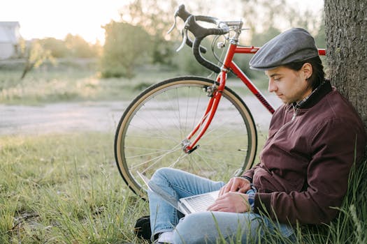 一个戴着平顶帽的年轻人正在公园里，靠着公路自行车，在笔记本电脑上工作
