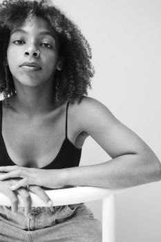 Striking black and white portrait of a woman with curly hair and nose piercing, leaning on a chair.