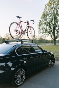 Red bicycle mounted on a black car with a roof rack, parked outdoors in a green park.