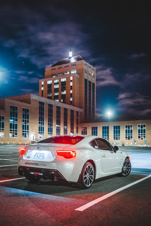 Stylish modern sports car with glowing headlights parked in parking lot near building against cloudy night sky