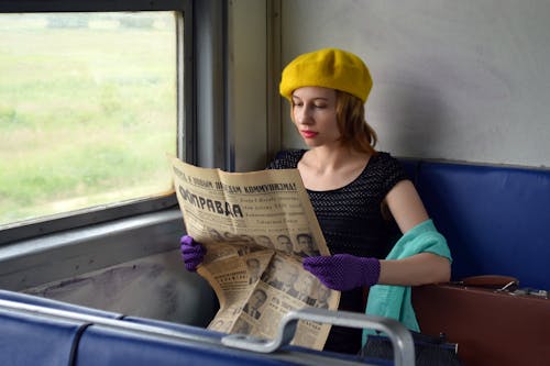 Free Concentrated young woman wearing elegant gloves and black blouse sitting in train with hand on suitcase and reading old Russian newspaper Stock Photo