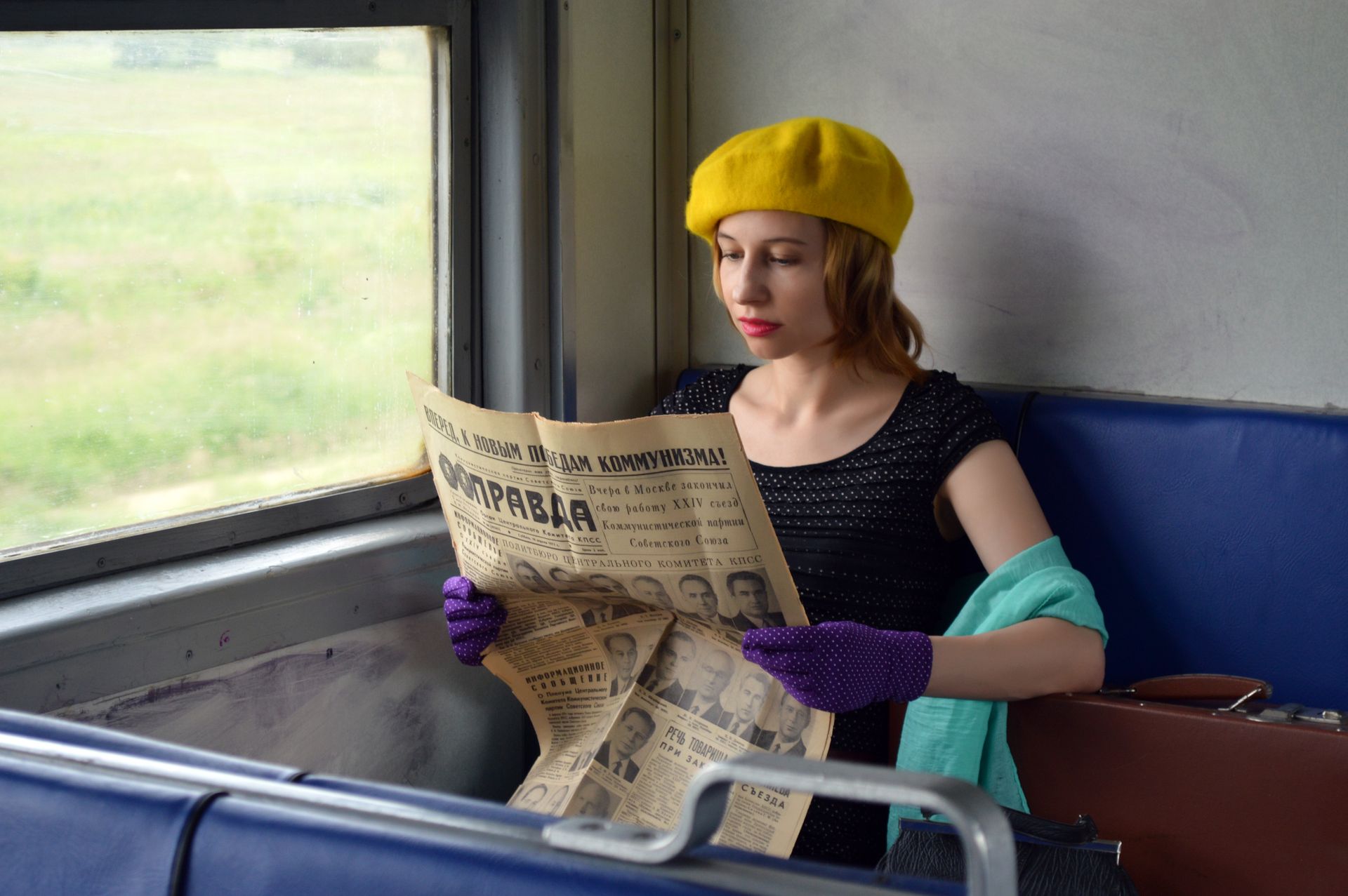 Concentrated young woman wearing elegant gloves and black blouse sitting in train with hand on suitcase and reading old Russian newspaper