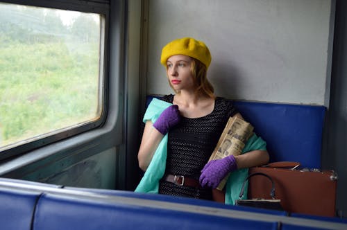 Free Calm young lady wearing elegant clothes sitting in train with newspaper and suitcase while looking out window in daytime Stock Photo