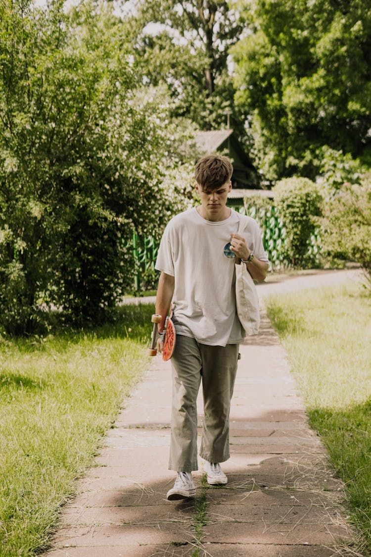 A Young Man Walking In A Park While Carrying A Skateboard