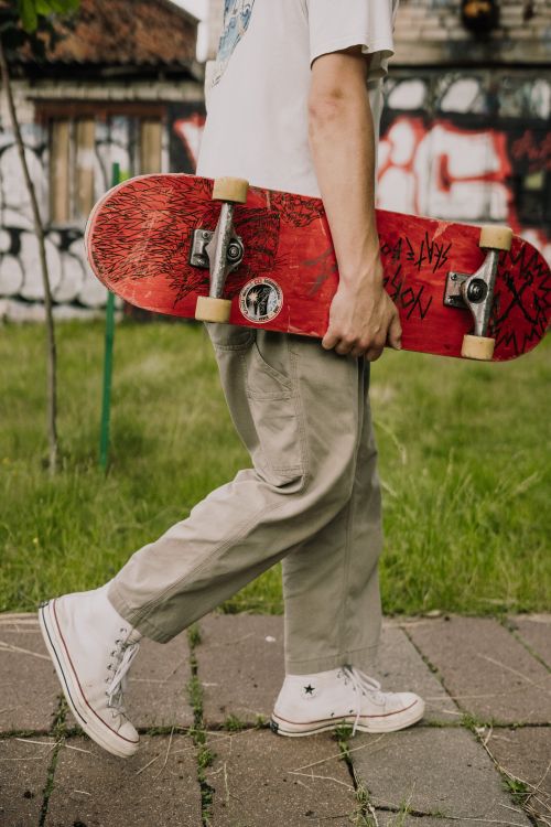 Free Close-up of a person in casual attire carrying a red skateboard while walking on a pavement outdoors. Stock Photo