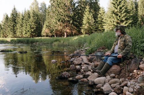 Free Senior man with beard fishing by a calm river surrounded by trees. Stock Photo