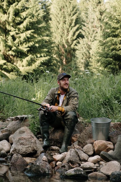 Free Bearded man fishing by a serene riverside amidst lush greenery, enjoying a summer day outdoors. Stock Photo