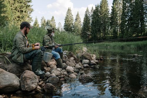 Free Two men enjoying a peaceful fishing trip by a forest riverbank, surrounded by nature. Stock Photo
