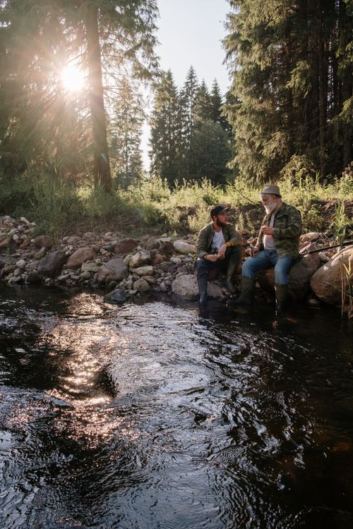 Free Two men sitting and fishing by a scenic riverbank in a lush forest during the day. Stock Photo