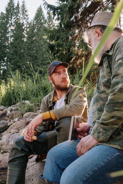 Free Two men sit by a riverbank, sharing a moment of leisure and connection in nature. Stock Photo