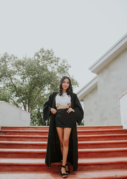Full body of young female in classic graduation clothes standing on stairs and looking at camera