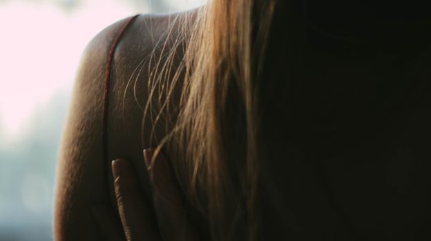 Back view of anonymous female with blond hair wearing cloth with thin straps while standing on blurred background with hand on back