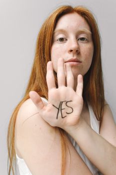Portrait of a woman with red hair showing 'yes' written on her palm in a bright setting.