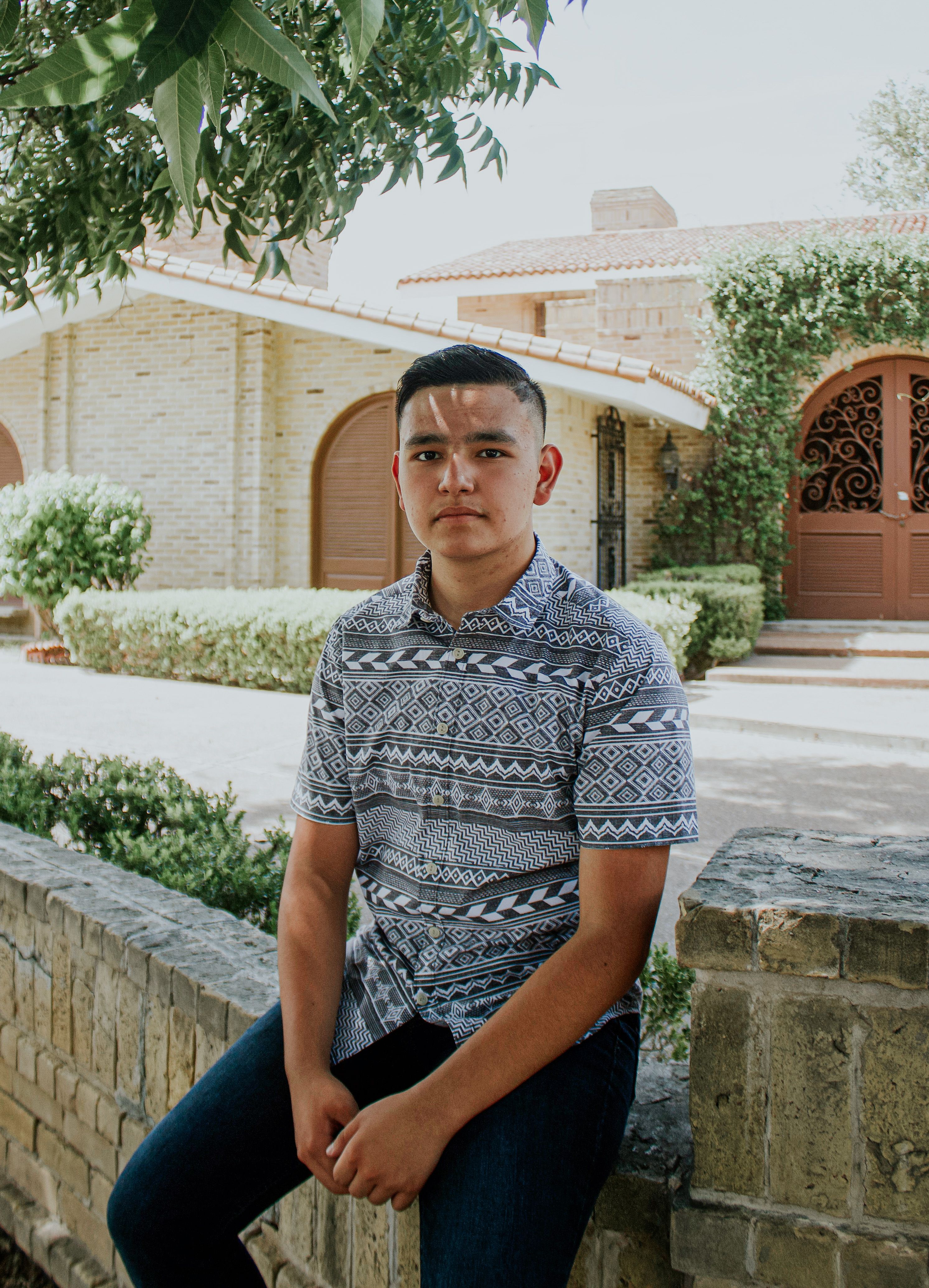Calm young male in summer shirt sitting on stone border near cozy house under green tree