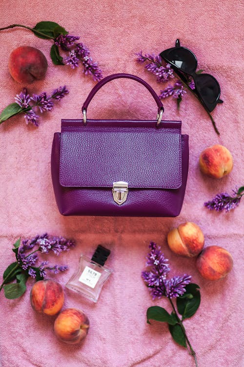 Free Top view composition of trendy violet handbag arranged on pink cloth amidst ripe peaches perfume bottle and stylish eyeglasses Stock Photo