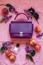 Top view composition of trendy violet handbag arranged on pink cloth amidst ripe peaches perfume bottle and stylish eyeglasses