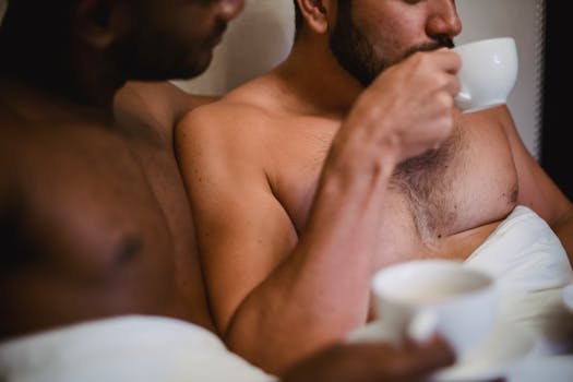 A tender moment with two shirtless men enjoying coffee in bed, symbolizing intimacy and togetherness.