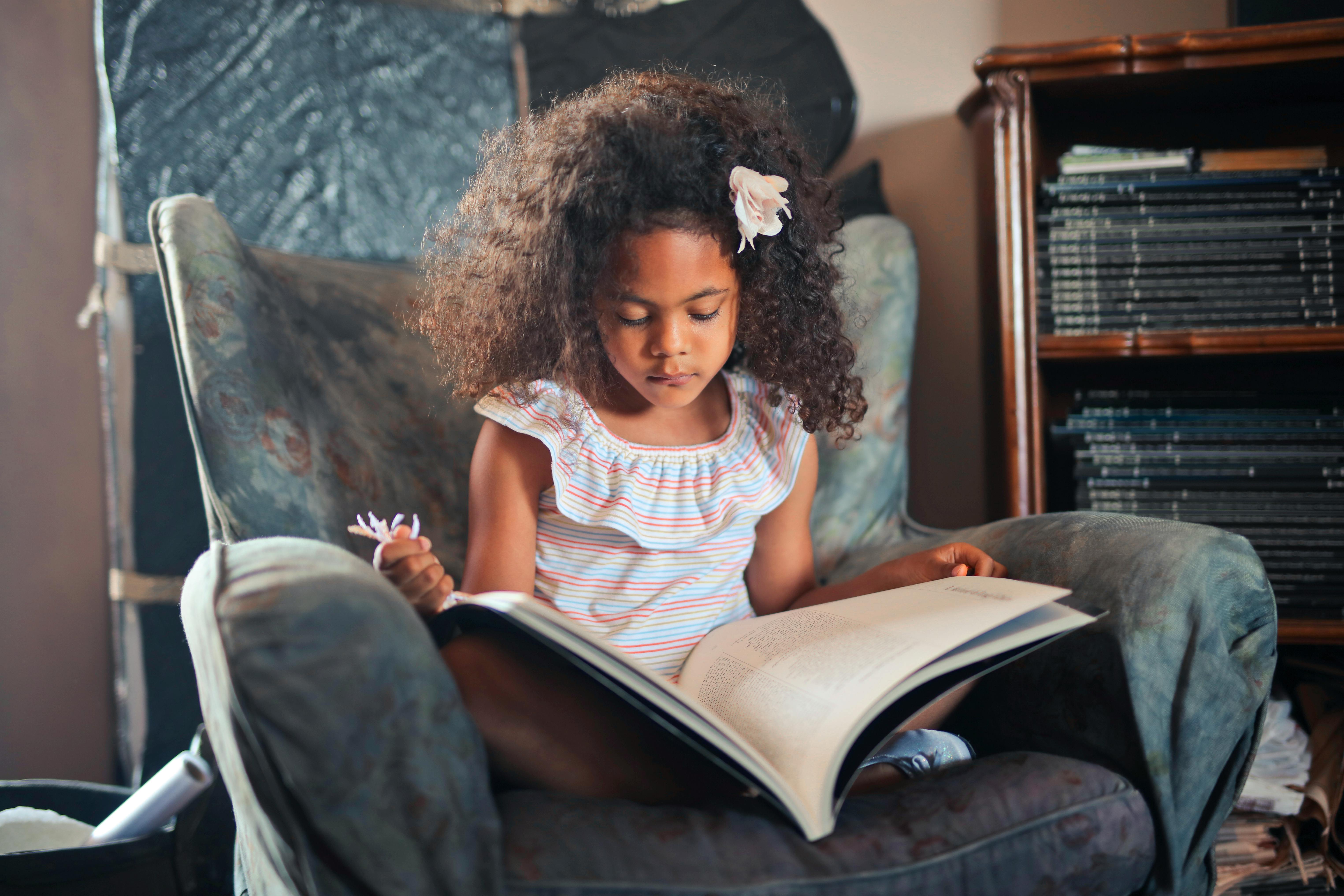Free A young girl reads an open book while sitting comfortably in an armchair indoors. Stock Photo