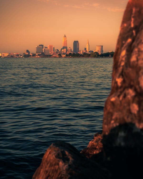 Picturesque view of calm ocean rippling near rock against sunset sky with skyscrapers in distance in evening time in coastal city