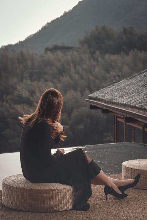Free Side view full body elegant female wearing classy black dress sitting on open terrace and enjoying views of rural house an lush tropical highlands Stock Photo