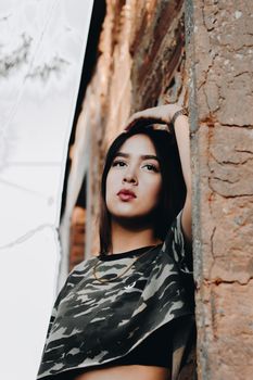 From below of dreamy young ethnic female in crop top touching head and looking away while leaning to old shabby cracked wall in daylight