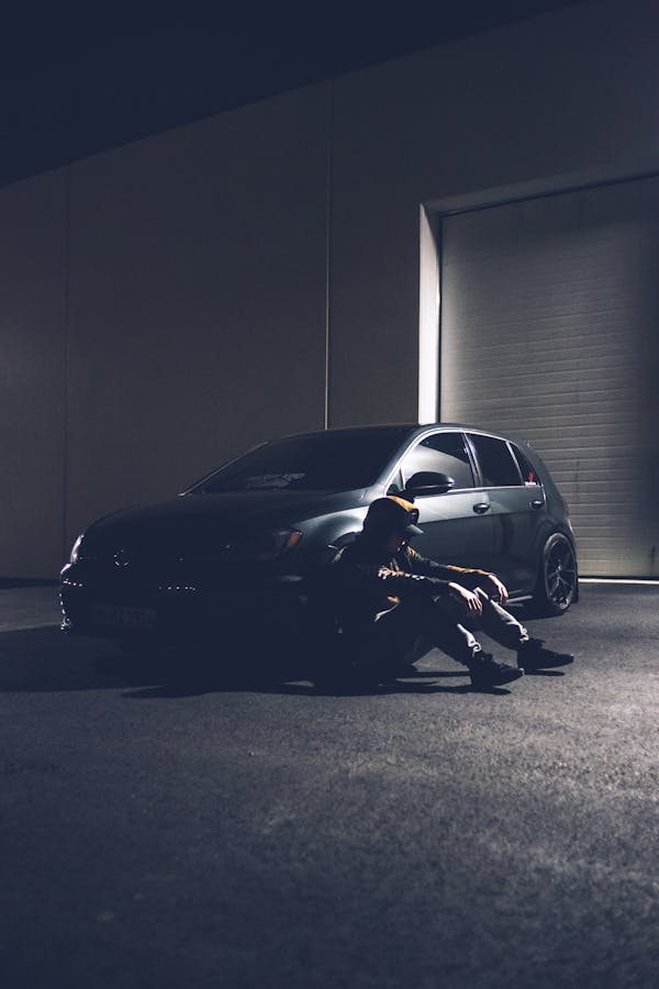 Side view of unrecognizable male in trendy outfit sitting on asphalt road near modern car parked in front of garage at night
