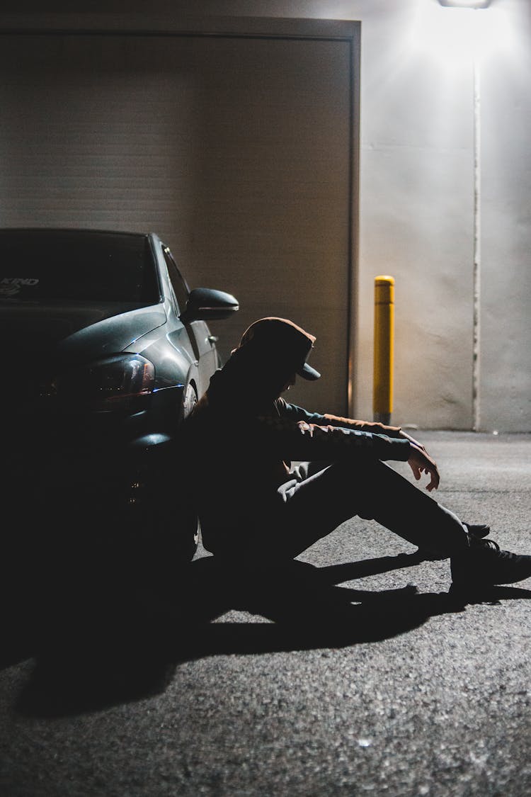 Anonymous Sad Man Sitting On Ground Near Parked Contemporary Car In Evening