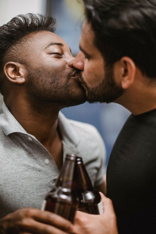 Free A close-up of an interracial gay couple sharing a romantic kiss with bottled drinks in hand, showcasing love and togetherness. Stock Photo