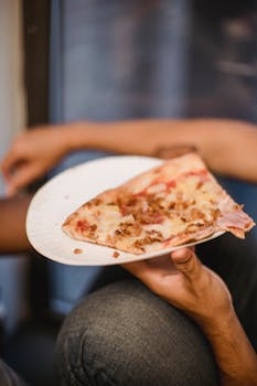 A close-up view of a hand holding a slice of pizza on a paper plate indoors.