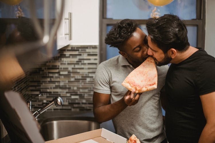 Two Men Biting A Slice Of Pizza