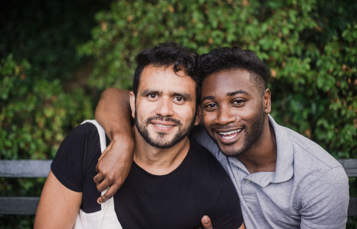 Two friends sitting on a bench outdoors, smiling and embracing warmly.