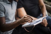 A Person in Gray Polo Shirt Sitting Next to a Person in Black Shirt while Flipping Pages of a Notebook