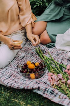 两个人正享受着宁静的户外环境，身旁是时尚的野餐布置，点缀着水果和鲜花