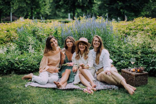 Women Sitting on a Picnic Blanket Holding Glasses of Wine While Smiling at the Camera