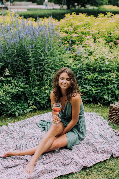 Woman Sitting on a Picnic Blanket near Green Plants with Flower while Holding a Glass of Wine
