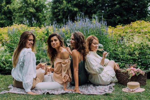 Women Sitting on a Picnic Blanket Near Green Plants with Flowers while Smiling at the Camera