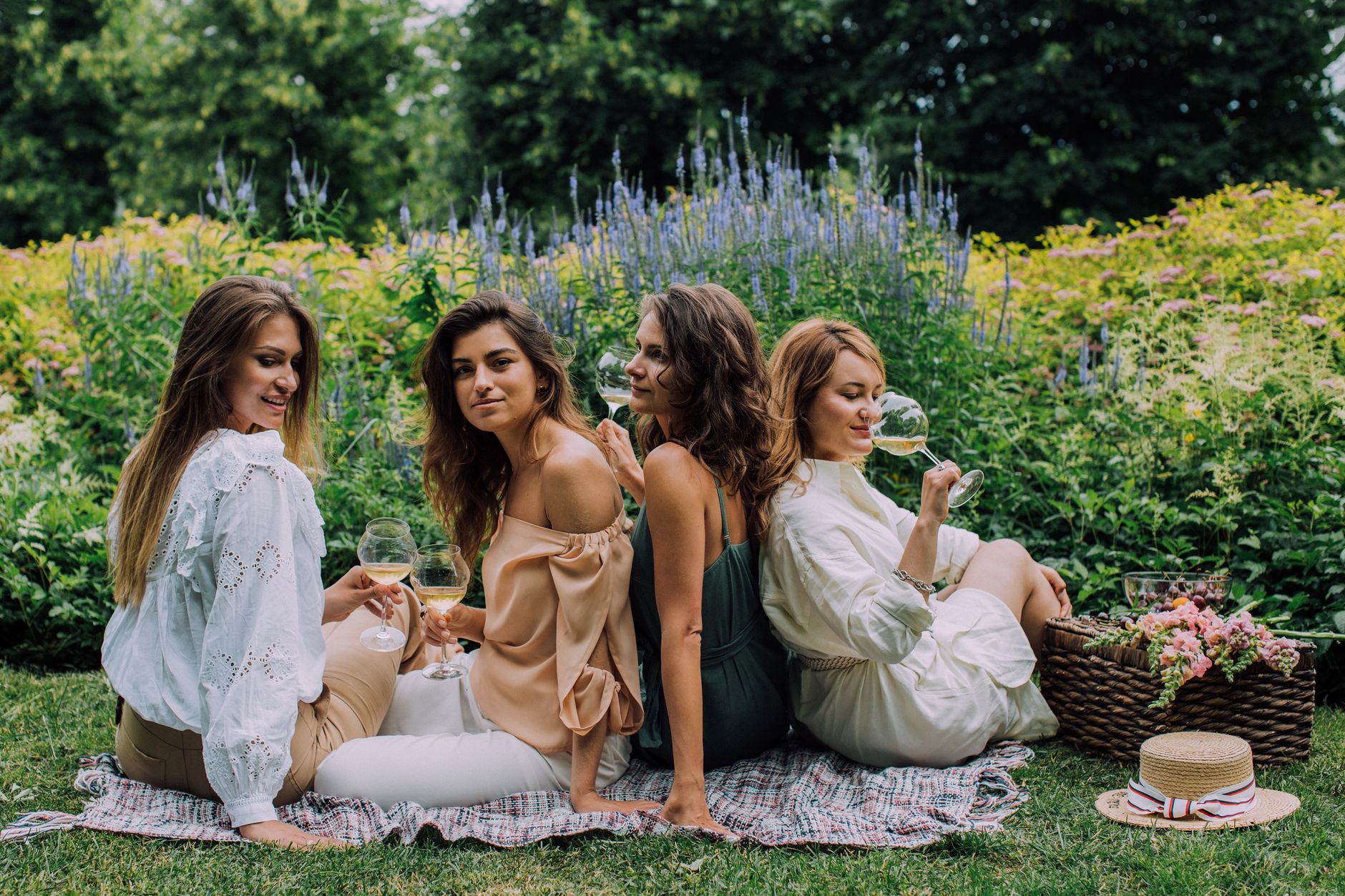 Four women enjoying a relaxed outdoor picnic with wine in a lush green garden setting.