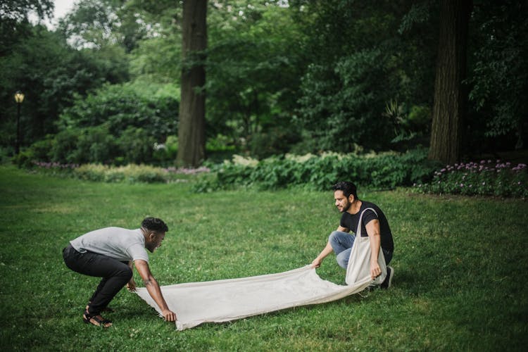 Men Laying Out The Blanket On The Grass Field Of A Forest Park