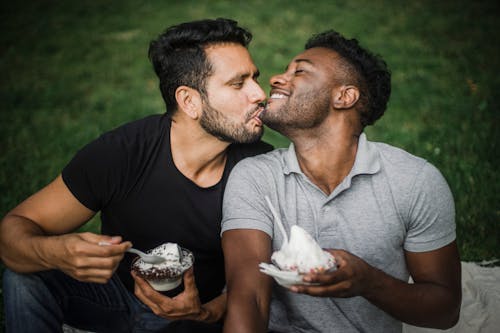 Man Kissing the Chin of the Man Sitting Beside Him while Holding Cups of Ice Cream