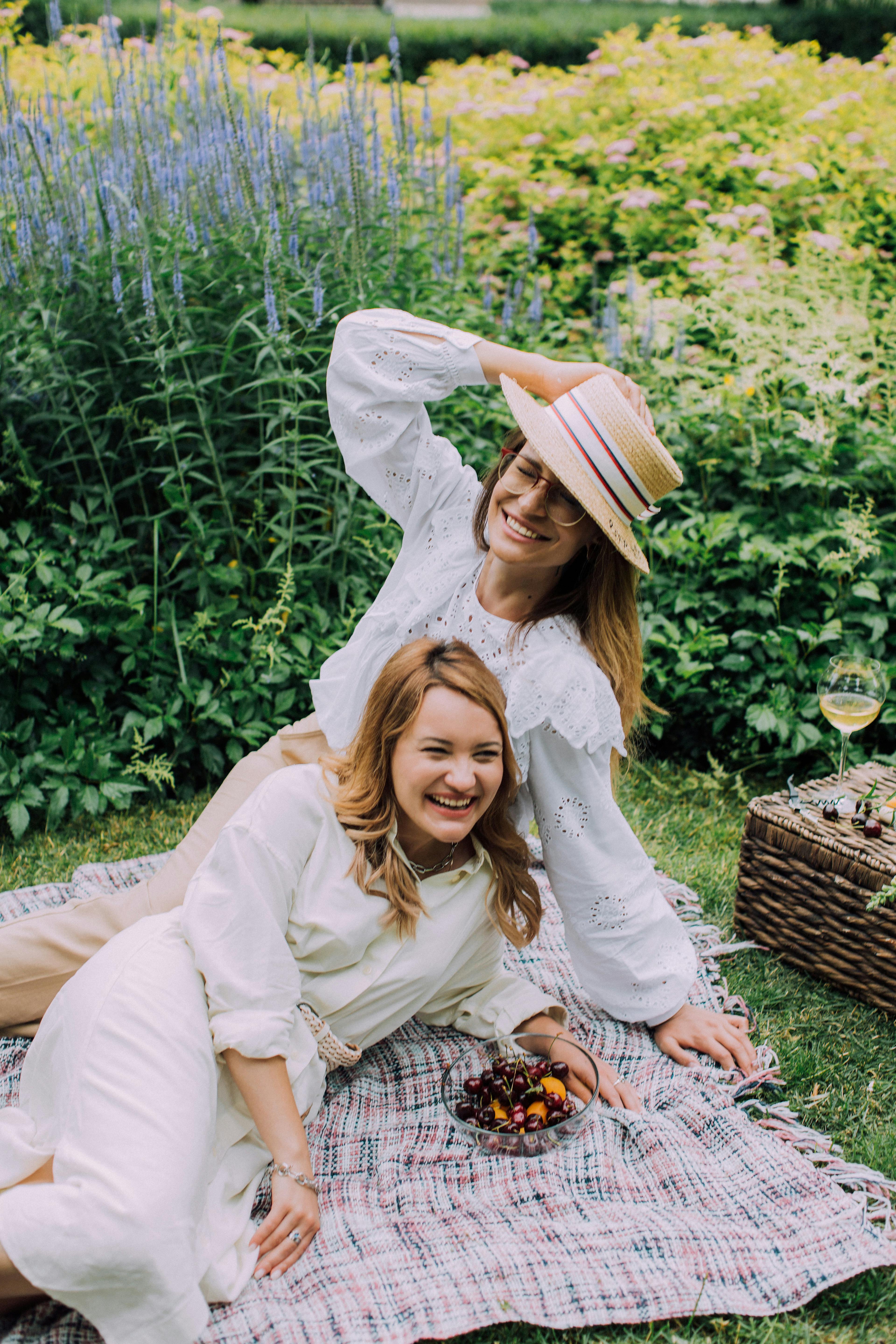Two women relaxing on a picnic blanket in a garden with a basket and wine, enjoying a sunny day.