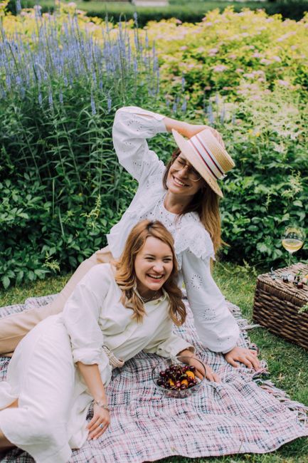 Two women relaxing on a picnic blanket in a garden with a basket and wine, enjoying a sunny day.