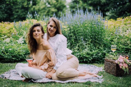 Free Two women sitting on a blanket in a lush garden with wine and flowers. Stock Photo