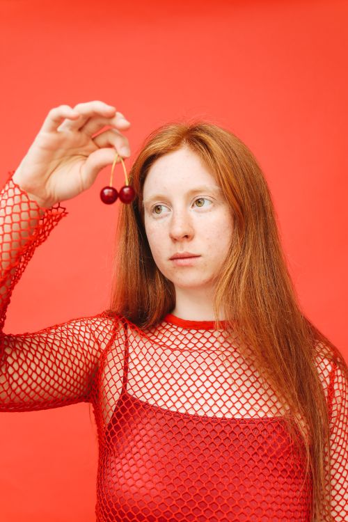 Free Portrait of a red-haired woman in a mesh top holding cherries against a bold red background. Stock Photo