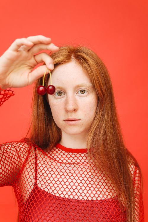 Free Portrait of a woman in a red fishnet top holding cherries against a red backdrop. Stock Photo