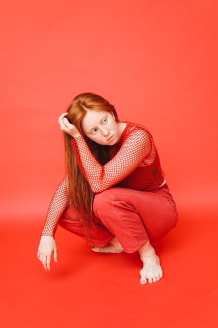 Caucasian woman with red hair posing barefoot in a studio against a red background.