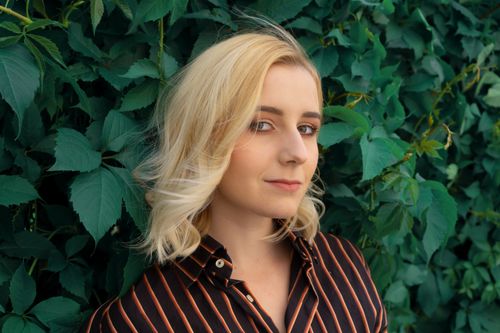 Free Young female with short blond hair and makeup in casual clothes standing against green leaves of shrub Stock Photo