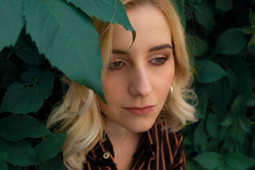 Free Pensive young female with blond hair and expressive makeup in striped shirt standing near green foliage of shrub Stock Photo