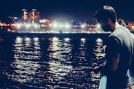 晚上，男子在码头钓鱼，城市灯光映在水面上